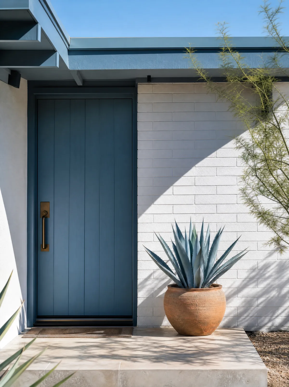 A blue-painted front door on a white brick mid-century Phoenix home, with a terracotta pot of blue agave and a crisp diagonal shadow