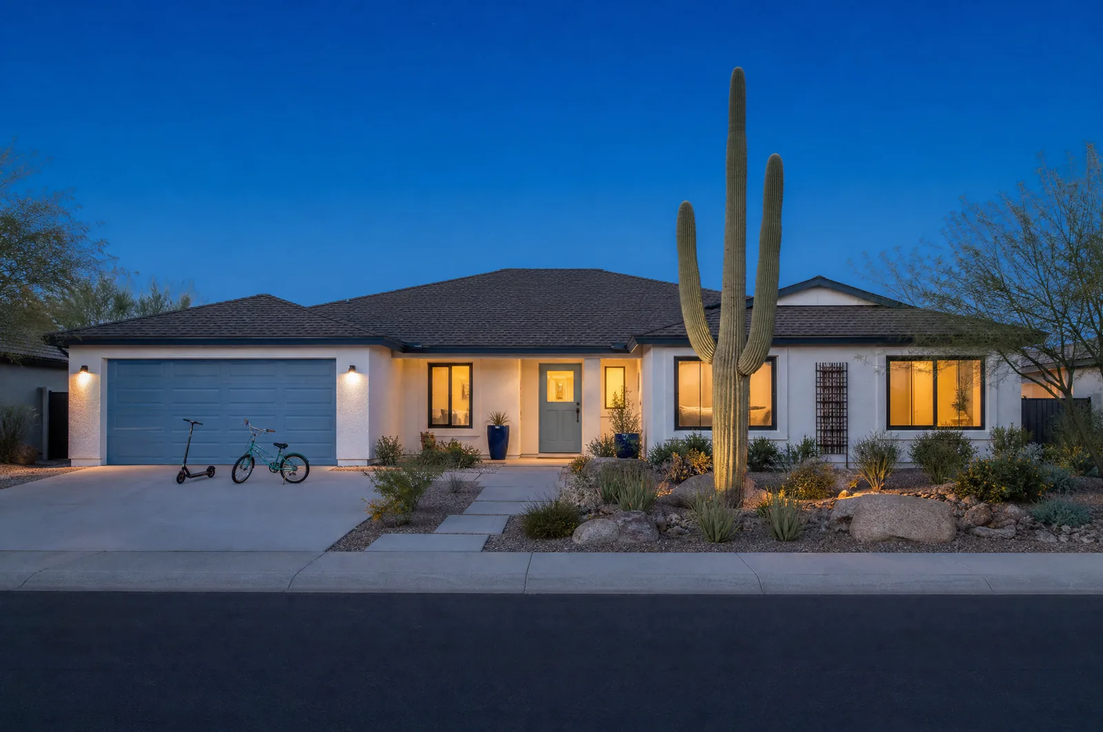 A single-storey family home in a Phoenix suburb at blue hour, with warm light in every window, a bicycle and a scooter by the garage and a saguaro in the front yard