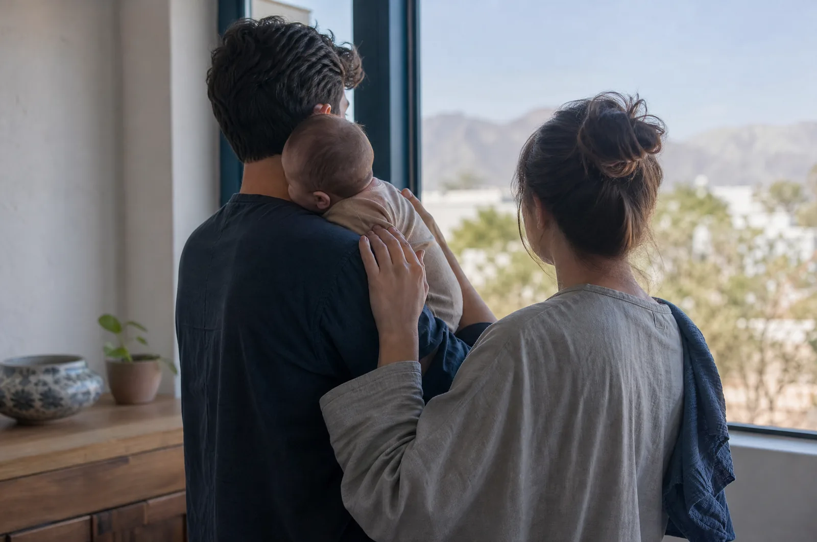 Two new parents standing at a window at home in Phoenix in soft morning light, one holding their infant against a shoulder, both seen from behind