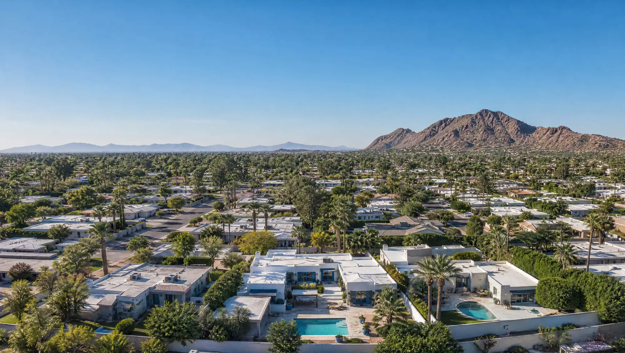 A low aerial view of a Phoenix neighborhood in clear morning light, with pale flat roofs, turquoise backyard pools, palms and citrus trees, and Camelback Mountain under a vast blue sky